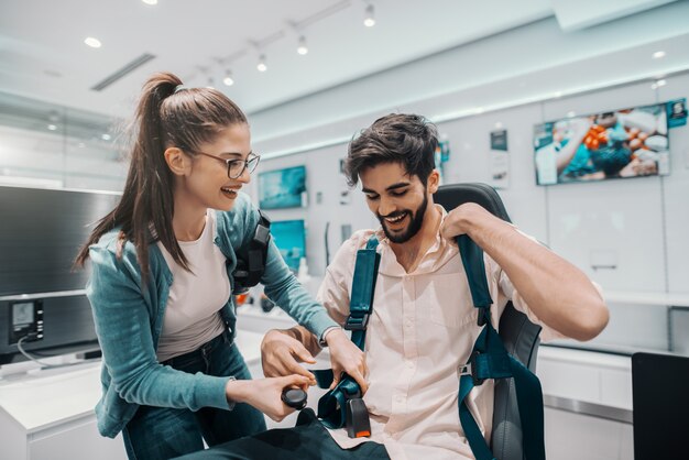 Smiling multicultural couple trying out virtual reality technology. Man sitting in chair while woman buckling belt. Tech store interior.