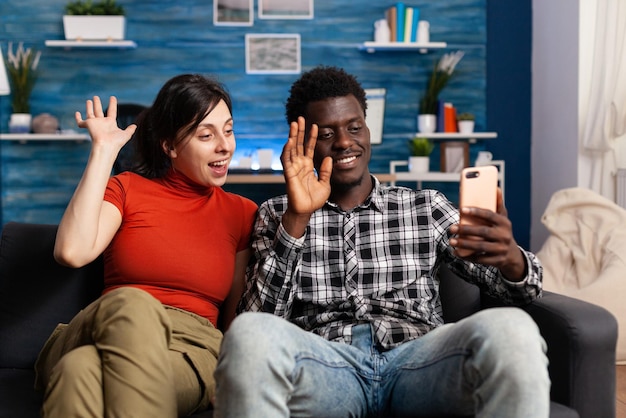 Улыбающаяся многонациональная пара машет смартфону во время видеоконференции дома. Молодые женатые люди обсуждают свою семью через онлайн-видеозвонок, сидя на диване в гостиной.