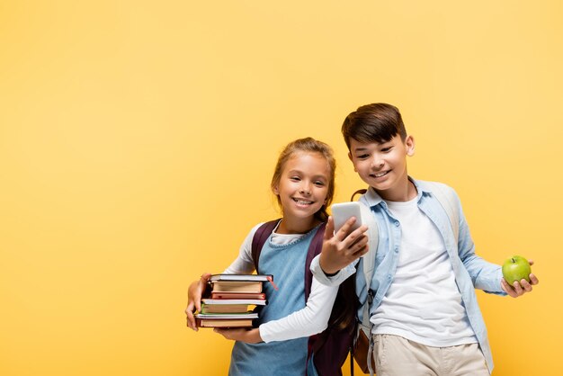 Smiling multiethnic pupils with books and apple using mobile phone isolated on yellowstock image