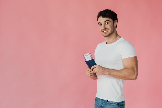 Photo smiling muscular man in white tshirt holding passport and air ticket isolated on pink