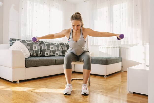 Smiling muscular woman in shape sitting on the chair at home and doing fitness exercises for arms with dumbbells.