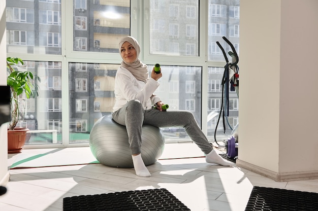 Smiling muslim girl in hijab exercising with dumbbells at home, looking away while sitting on a fit ball