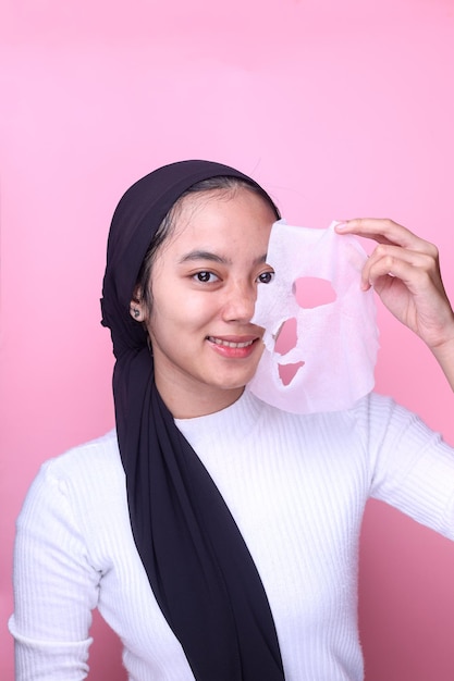 smiling muslim teen girl doing skin care routine, peel off moisturizer sheet mask on pink background