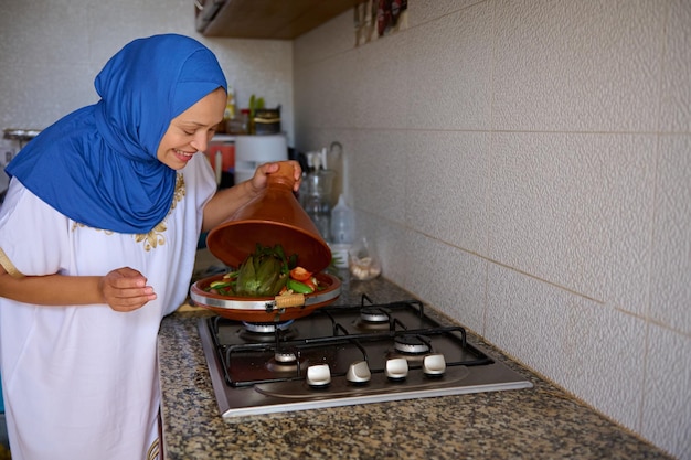 smiling muslim woman in blue hijab opening lid of a clay dish while cooking veggies in moroccan tagine at home kitchen