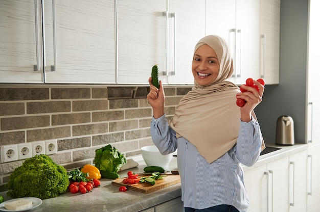 Sorridente donna musulmana in hijab mostra un cetriolo e pomodori alla telecamera mentre prepara un'insalata vegana in cucina.