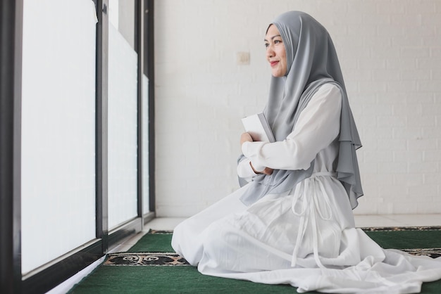 Smiling muslim woman holding holy book Quran on chest looking away while sitting on carpet inside m