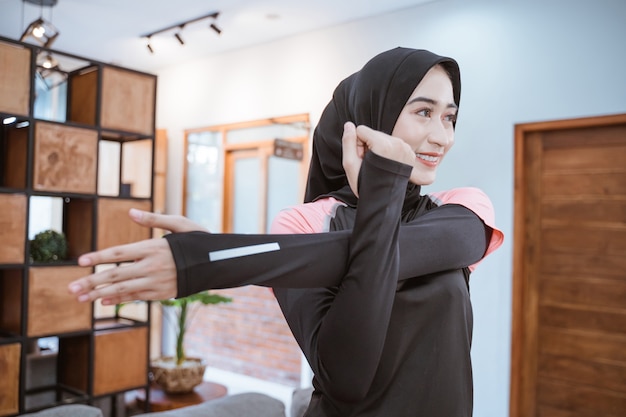 Smiling muslim woman in sports clothes stands stretching arms in the house