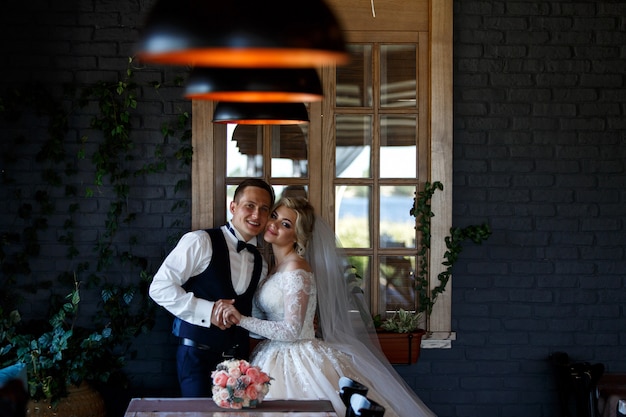 Smiling newlyweds hugging near the window indoor. portrait of newlyweds in stylish hotel room. wedding couple in a room with a stylish interior with lamps. wedding day