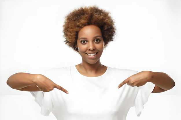 Photo smiling nice african american woman pointing at blank white tshirt with both index fingers copy space for your advertising