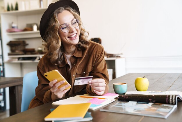 Foto bella donna sorridente con cappello che usa il cellulare e tiene la carta di credito mentre studia a casa