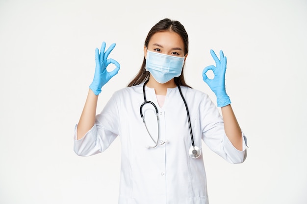 Smiling nurse female asian doctor in rubber gloves medical face mask shows okay ok signs stands agai...