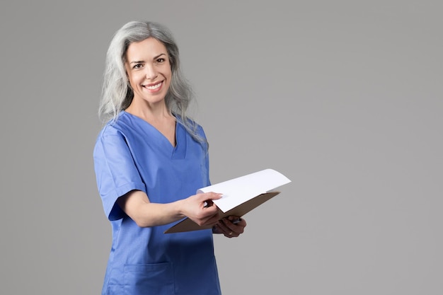 Улыбающаяся медсестра с медицинской картой позирует на сером фоне