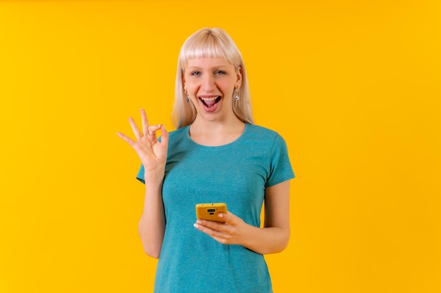 Smiling ok gesture with phone blonde caucasian girl on yellow studio background