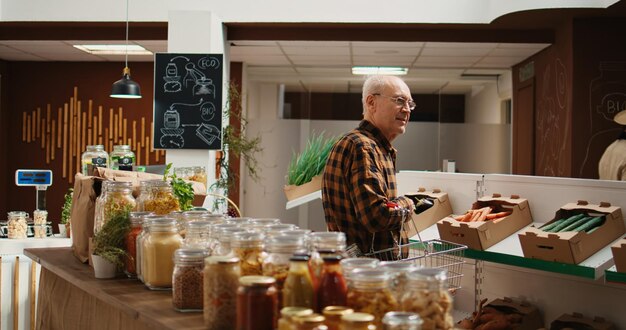 폐기물 제로 슈퍼마켓 진열대에서 식품 저장실 필수품을 찾고 있는 웃고 있는 늙은 고객. 환경 친화적인 지역 식료품점에서 재사용 가능한 항아리에 유기농 식품을 구입하는 행복한 노인