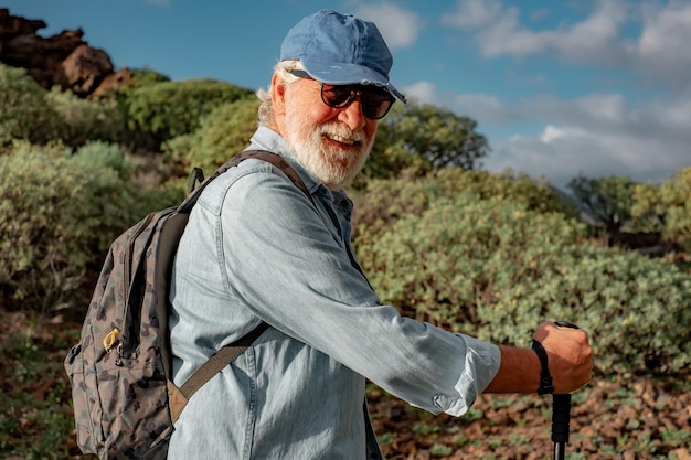 Улыбающийся пожилой мужчина наслаждается походами по сельской местности в рюкзаке и шляпе Пожилой кавказец с тростью смотрит в камеру