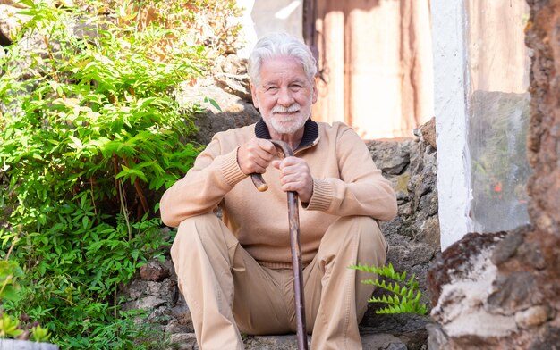 Smiling old senior man in winter sweater outdoors of a rural house leaning on walking stick. Caucasian man with beard and white hair