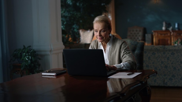 Smiling old woman reading good news on laptop computer in luxury interior Aged lady working computer in vintage cabinet Senior businesswoman using pc on home workplace