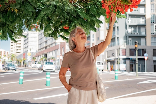 Smiling old woman wearing eyeglasses walking in city street under a tree to touch the red flowers