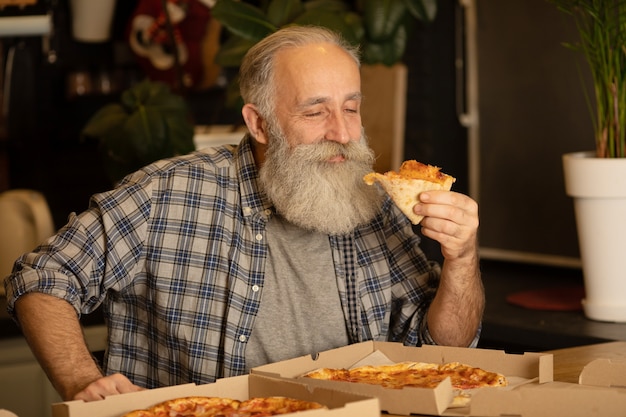 Smiling older man eating pizza slice sitting at living room. Bearded senior man eating italian food.