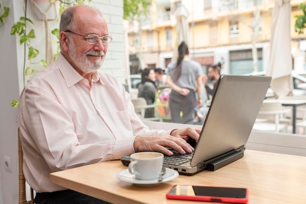 A smiling older man with eyeglasses sitting in the coffee shop bar drinking coffee and working on his laptop