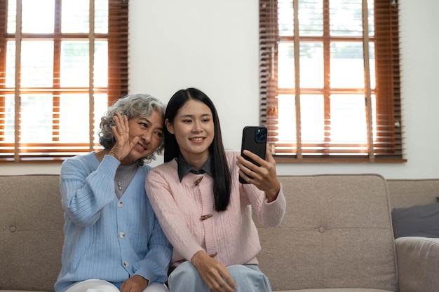 웃는 나이 많은 어머니와 성인 딸이 함께 집에 있는 아늑한 소파에 앉아 전화를 사용하고 있습니다.