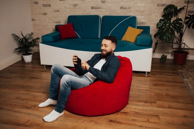 Smiling optimist carefree bearded man in casual clothes sit indoor modern interior and using his smart phone while sitting