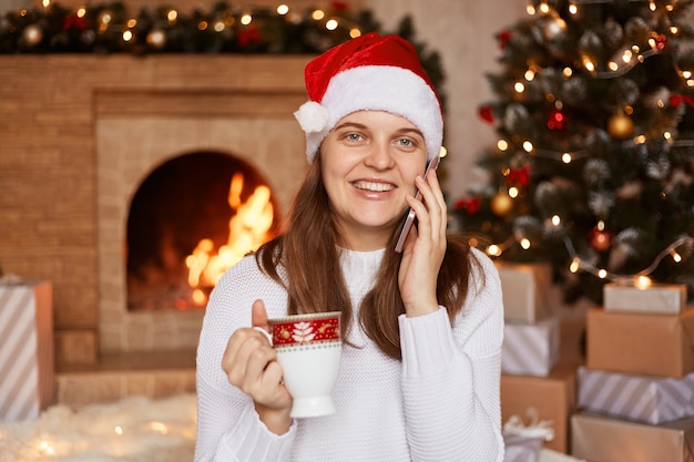 Smiling optimistic young adult woman wearing white sweater and santa claus hat, posing near fireplace and xmas tree in festive living room and talking via smart phone.