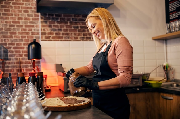 A smiling pancake house worker is preparing crepes and putting chocolate fill on it