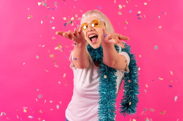 Smiling at a party throwing confetti in sunglasses blonde caucasian girl on pink background studio