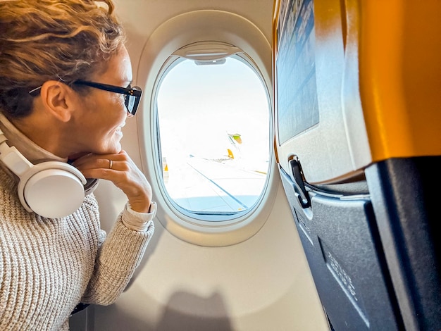 Photo smiling passenger with bluetooth headphones looking out of the airplane porthole middleaged woman sitting by the window relaxing while waiting to arrive