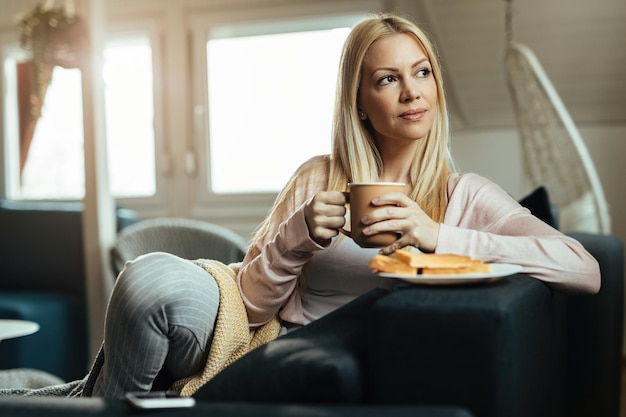 Smiling pensive woman relaxing on the sofa and enjoying in cup of coffee in the living room