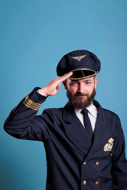 Photo smiling plane pilot saluting, wearing uniform and hat front view portrait, airplane captain looking at camera. civil aviator with badge on jacket, aircrew member on blue background