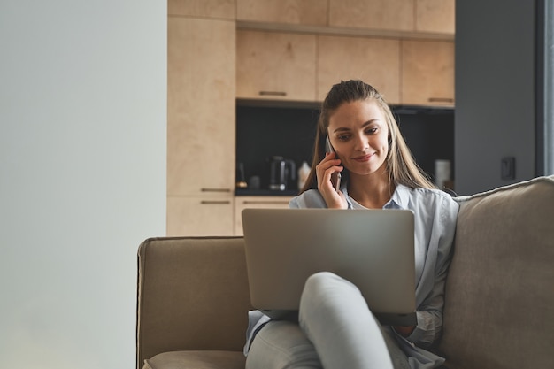 Smiling pleasant modern young blonde Caucasian female freelancer with a laptop sitting on the couch