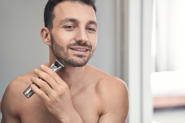 Smiling pleasant young male preparing to use an electric razor to shave his beard in the morning