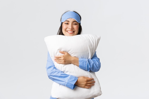 Улыбающаяся довольная азиатская девушка в спальной маске и пижаме обнимает мягкую и удобную подушку с