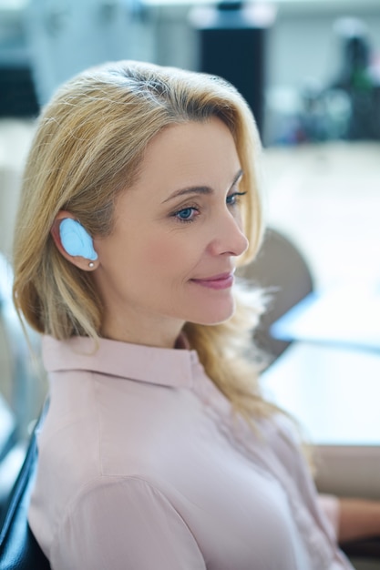 Smiling pleased beautiful blonde woman with a device inserted into the ear sitting at the doctors office