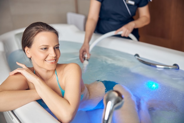 Smiling pleased beautiful young dark-haired female enjoying an underwater shower massage in a spa salon