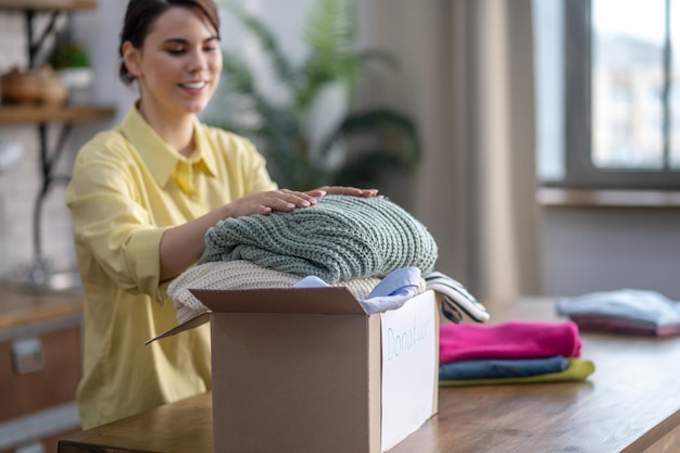Smiling pleased concentrated attractive young woman putting two folded knitwear pullovers in the cardboard box
