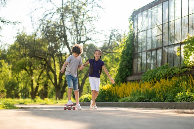 Smiling pleased dark-haired young lady helping a focused beginner skateboarder to step on the board