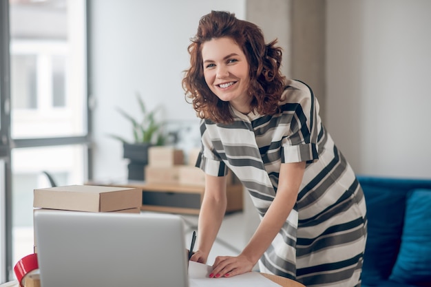 Smiling pleased employee with a ball-point pen in her hand leaned over the office desk looking ahead