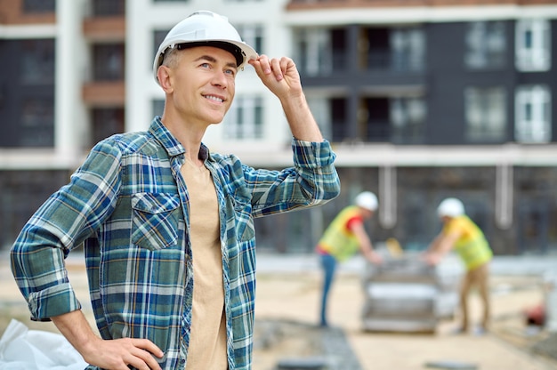 Улыбающийся довольный красивый инженер-строитель трогает его каску