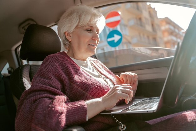 Sorridente compiaciuta signora dai capelli grigi seduta nel salone dell'auto e guardando lo schermo del laptop