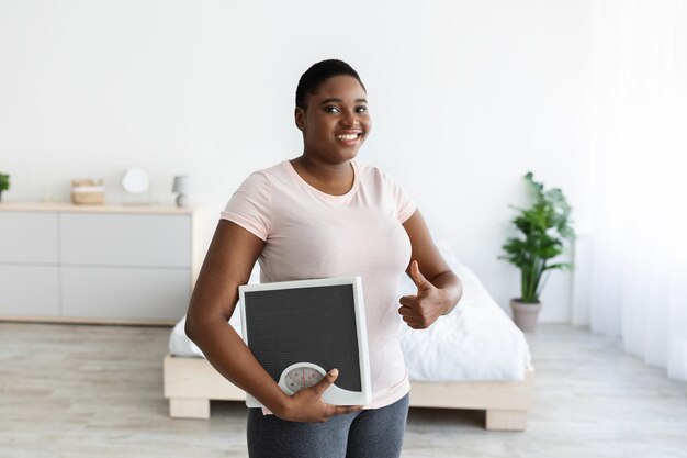 Smiling plus size black woman holding scales showing thumb up gesture happy with result of her