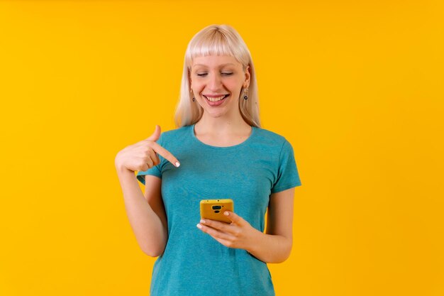 Smiling pointing at the phone blonde caucasian girl in studio on yellow background