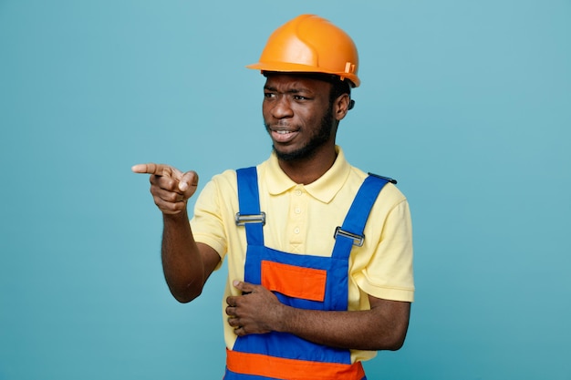 Smiling points at side young african american builder in uniform isolated on blue background