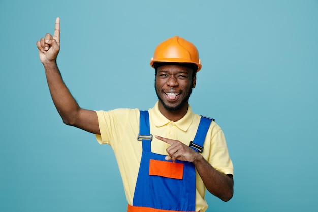 Smiling points at side young african american builder in uniform isolated on blue background