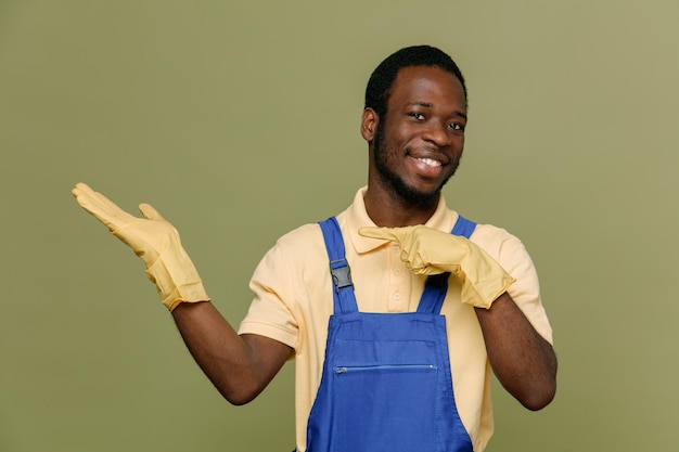 Punti sorridenti a lato giovane pulitore afroamericano maschio in uniforme con guanti isolati su sfondo verde