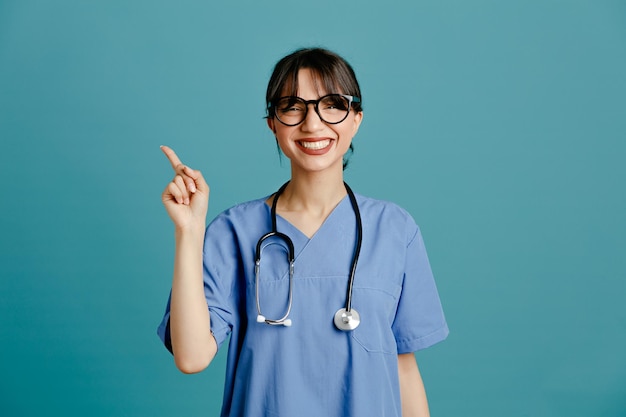 Smiling points at side young female doctor wearing uniform fith stethoscope isolated on blue background