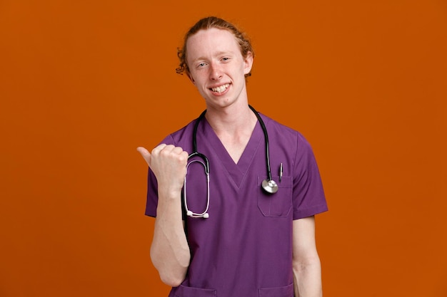 Smiling points at side young male doctor wearing uniform with stethoscope isolated on orange background