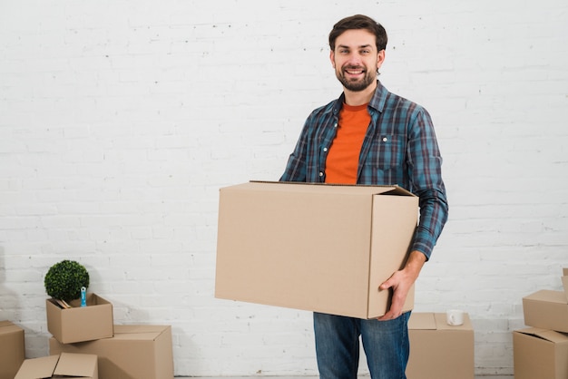 Photo smiling portrait of a young man carrying the cardboard box standing against white wall